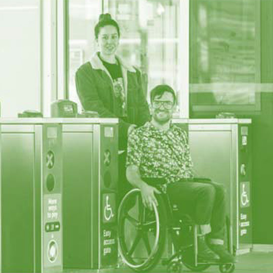 A smiling person in a wheelchair is being assisted through a railway station ticket gate by a standing person.